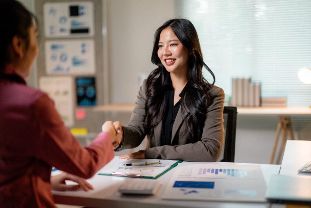 Two women sit across from each other at an office desk, shaking hands. Charts and documents are spread on the desk, with office supplies and shelves in the background.