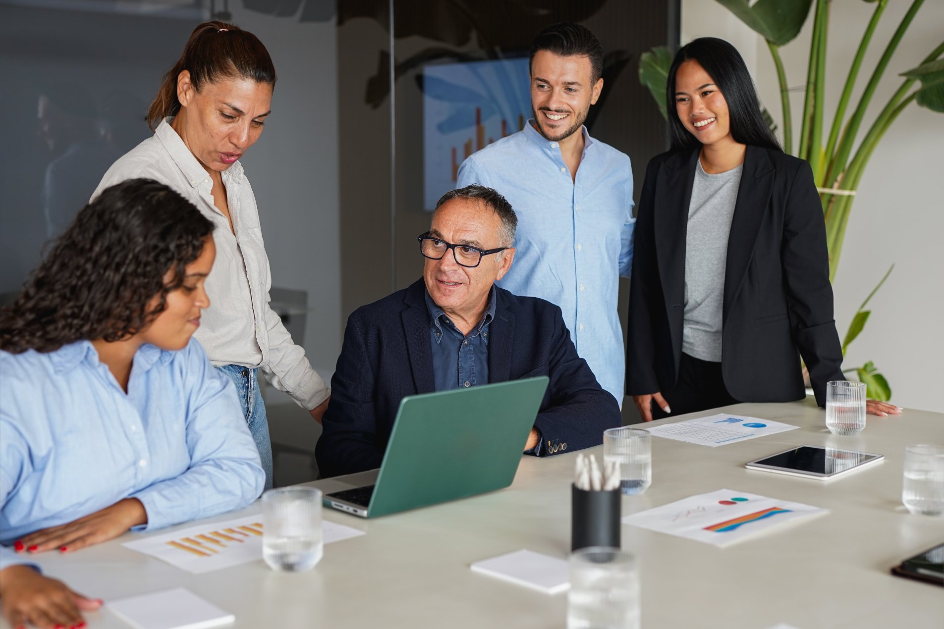 Five people gather around a table with a laptop, papers, and water glasses, having a discussion in a modern office setting.