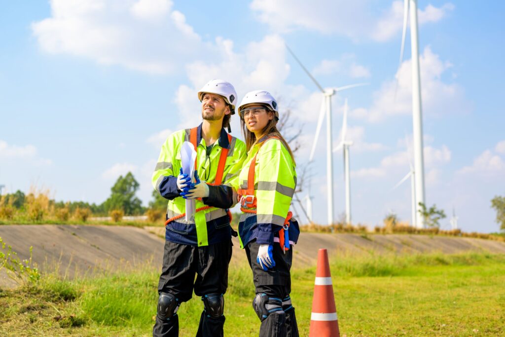 Two engineers in safety gear and helmets stand near a traffic cone outdoors, holding blueprints with wind turbines in the background.