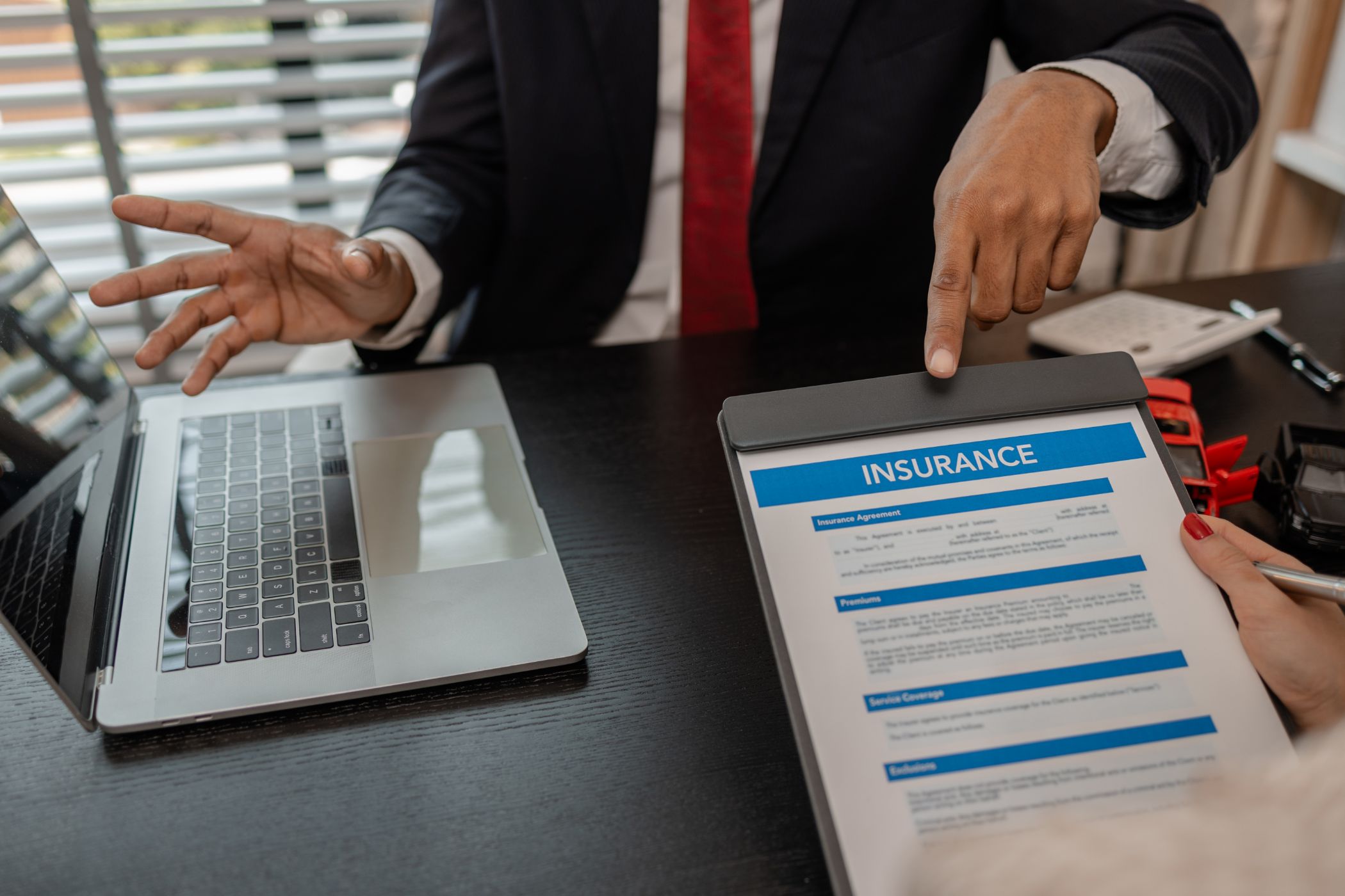Two people sit at a desk with a laptop, one person holding and reviewing an insurance document while the other gestures and points at the document.
