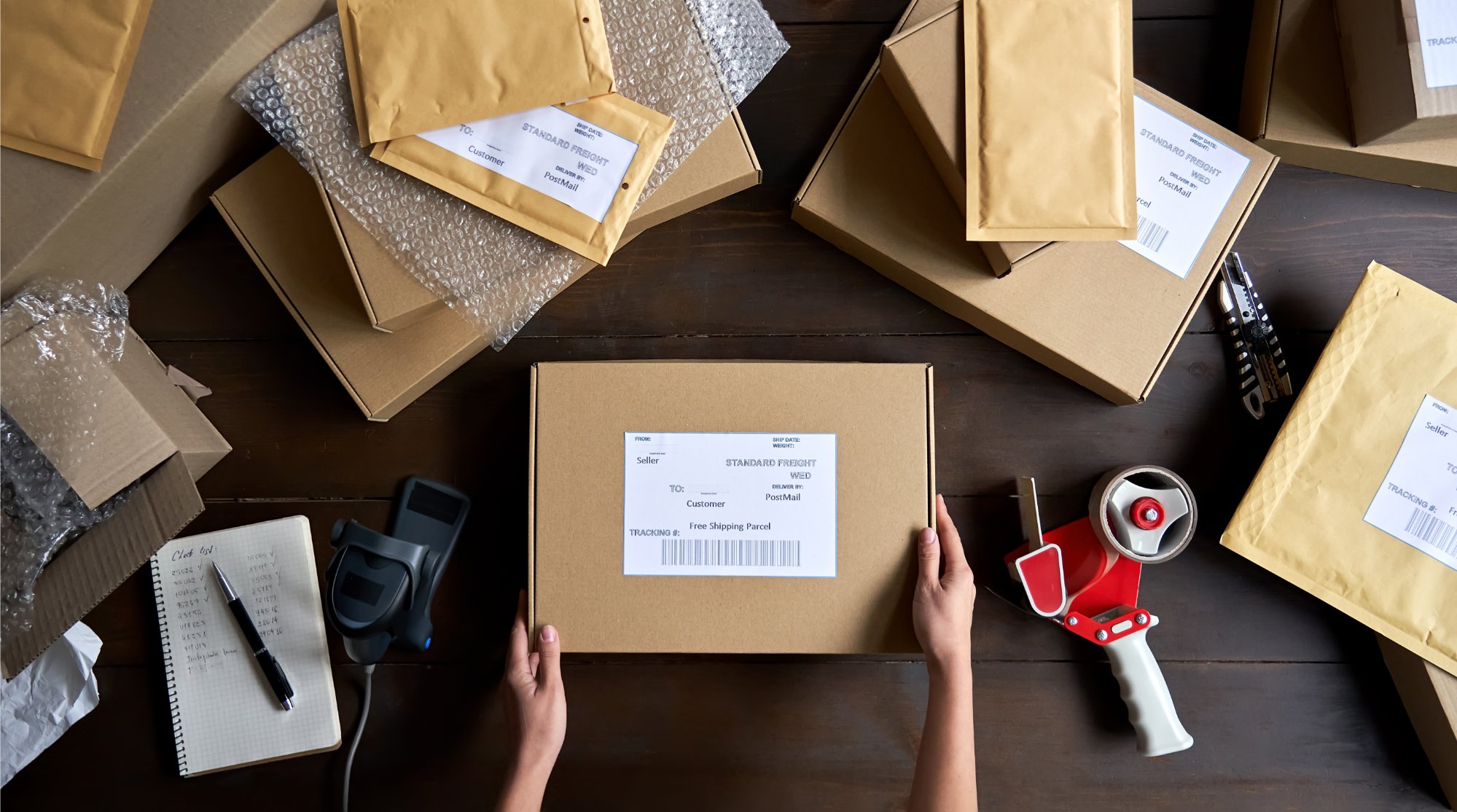 A person prepares packages for shipping on a wooden table, surrounded by boxes, envelopes, a tape dispenser, bubble wrap, and a notepad.