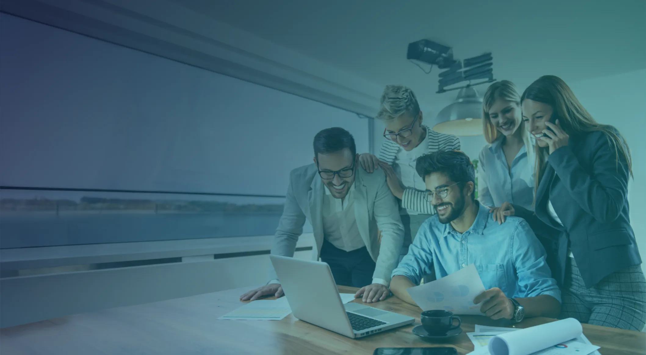 Five people gather around a table, looking at a laptop and documents, appearing to collaborate and discuss work in a modern office setting.